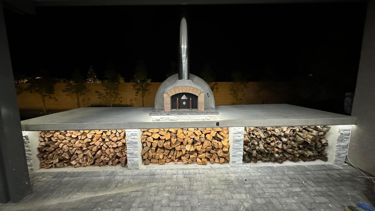 Brett’s outdoor masonry pizza oven, with a metal chimney and stacked firewood underneath.